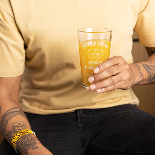 A man with tattoos holding our classic "American Conservative" Donald Trump pint glass filled with orange juice.